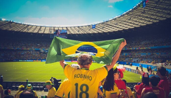 copa-camisa-amarela A imagem mostra um torcedor de costas, levantando a bandeira do Brasil sobre a cabeça, em um estádio. Ele usa a camisa amarela número 18 com o nome Robinho.
